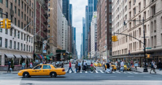 Busy city street in Manhattan with pedestrians crossing a wide crosswalk and yellow taxis parked along the curb or in traffic.