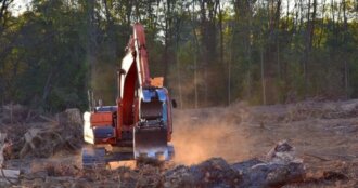 Excavator with red arm at a dusty construction site, clearing land with trees in the background